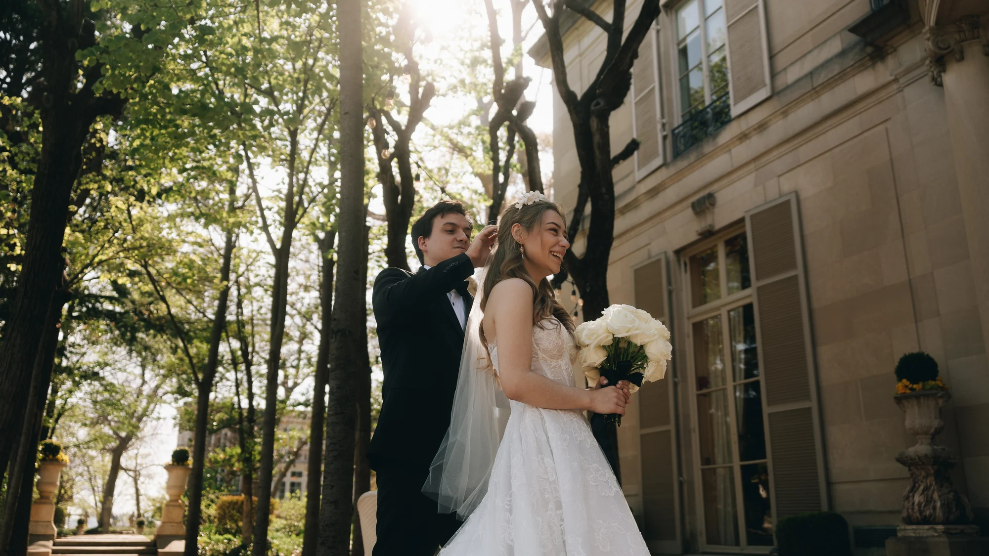 The bride and groom under bare spring trees beside the stone house