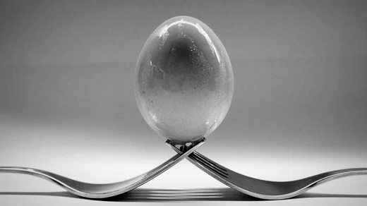 An egg balanced on the crossed tines of two forks, in black and white