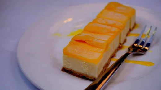 A rectangular mango cheesecake sliced into cubes, a dessert fork resting beside it on a white plate