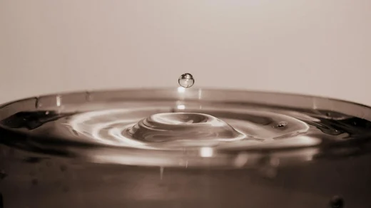 A single droplet suspended above the rippling surface of a full pot, caught in sepia