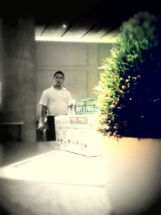 A man standing behind boxes at a supplement display, the frame desaturated except one green plant