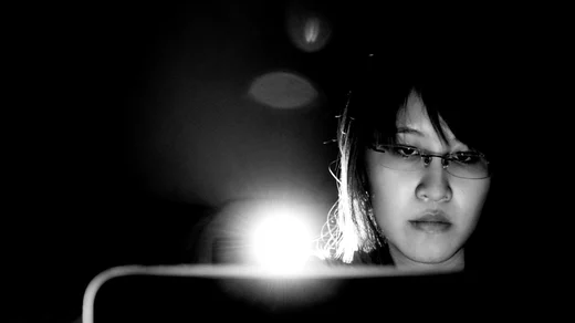 A woman in glasses lit only by her laptop screen, in black and white