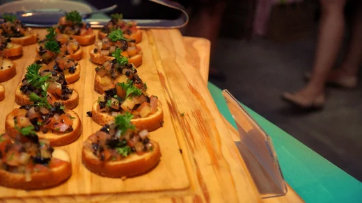 Rows of bruschetta garnished with parsley on a wooden board, guests blurred beyond the table
