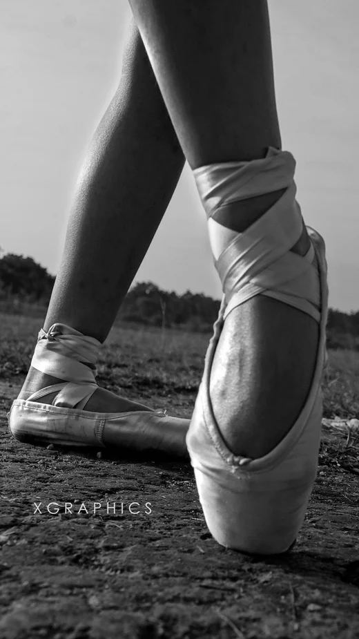 A dancer's crossed feet in ribboned pointe shoes, en pointe on bare ground, in black and white