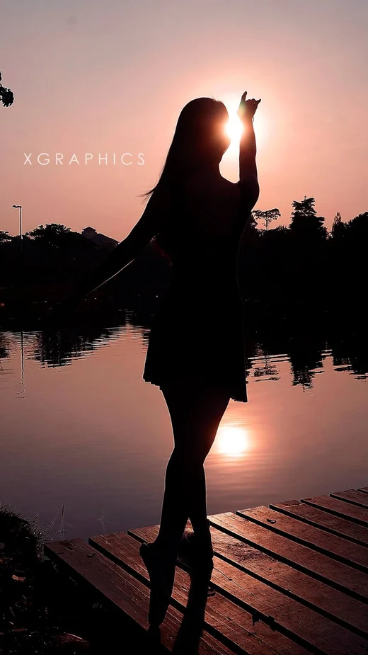 A woman silhouetted on a lakeside dock at sunset, one arm raised toward the sun