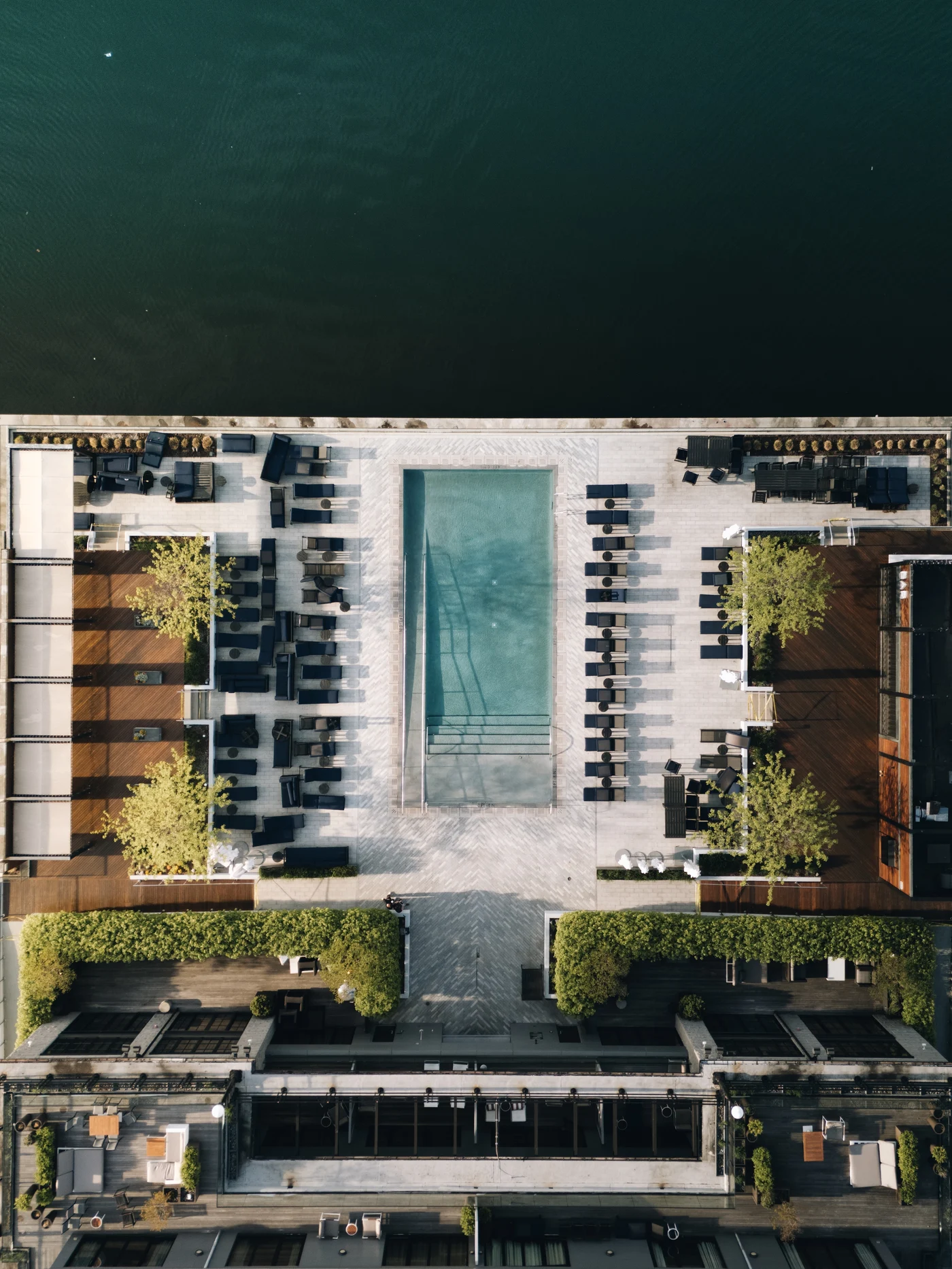 Directly overhead: a long pool between two ranks of loungers, planting and cabanas framing it, the water of the harbour along the top edge