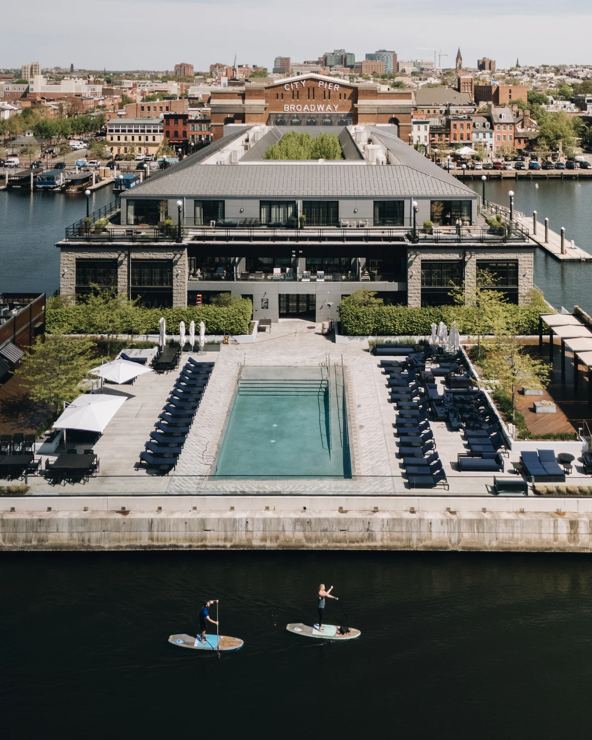 From out on the water, the low waterfront building with its pool and rows of loungers, city rooftops behind, two paddleboarders crossing the foreground