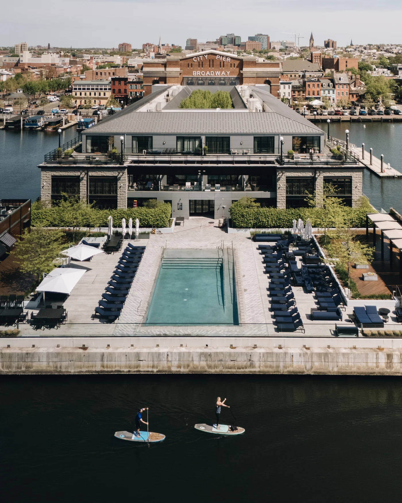 From out on the water, the low waterfront building with its pool and rows of loungers, city rooftops behind, two paddleboarders crossing the foreground