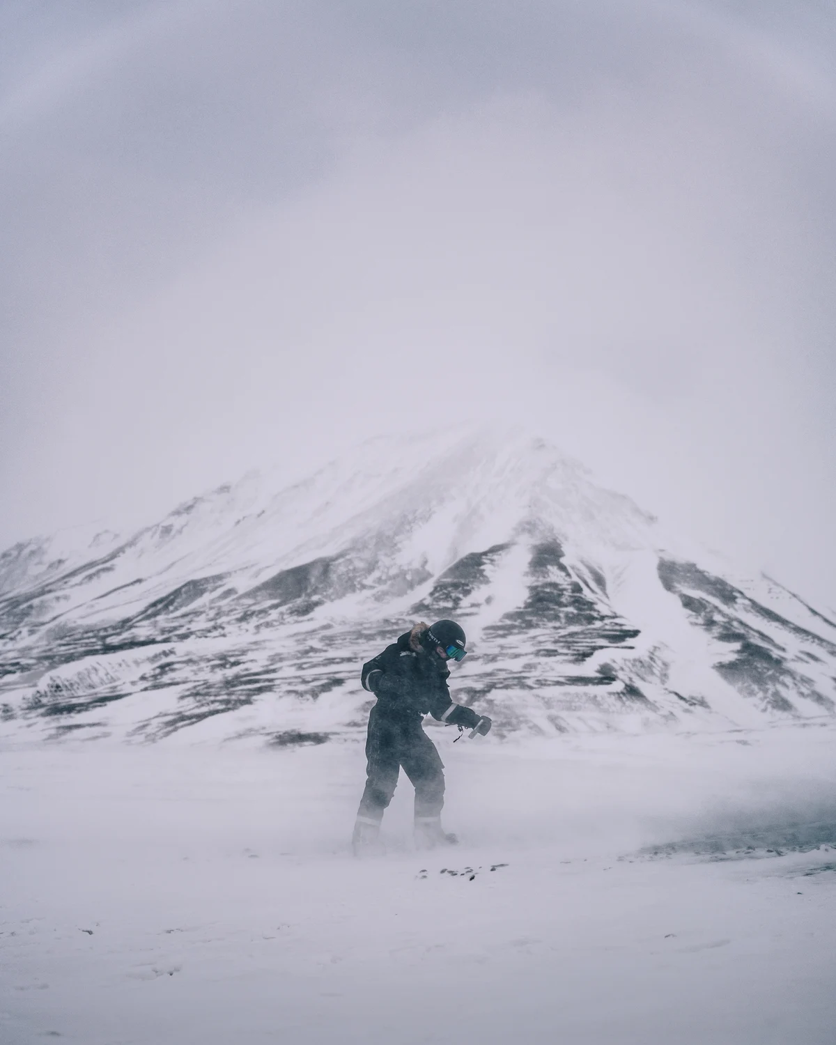 A guide crossing a frozen plain in blowing spindrift, a striped snow mountain rising into cloud behind