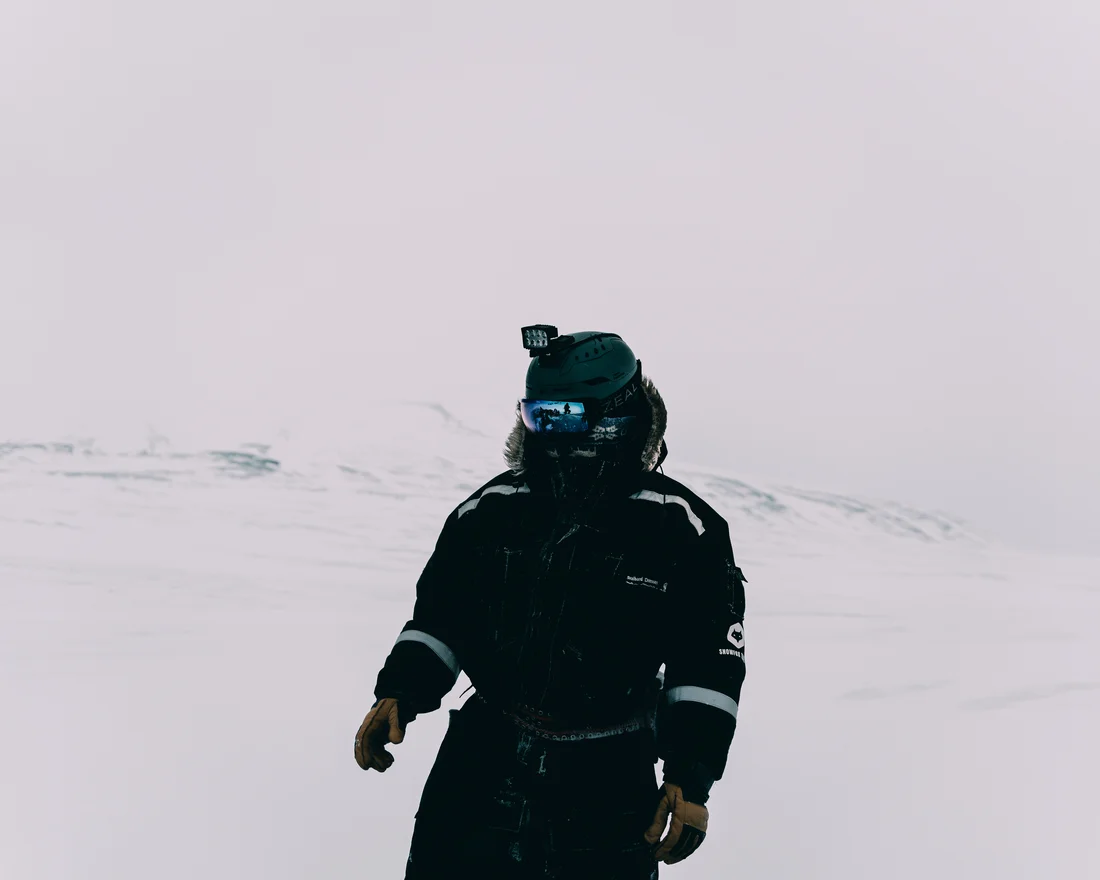 A guest in a black expedition suit and helmet alone on an empty white plain, the rest of the group reflected small in the mirrored goggles