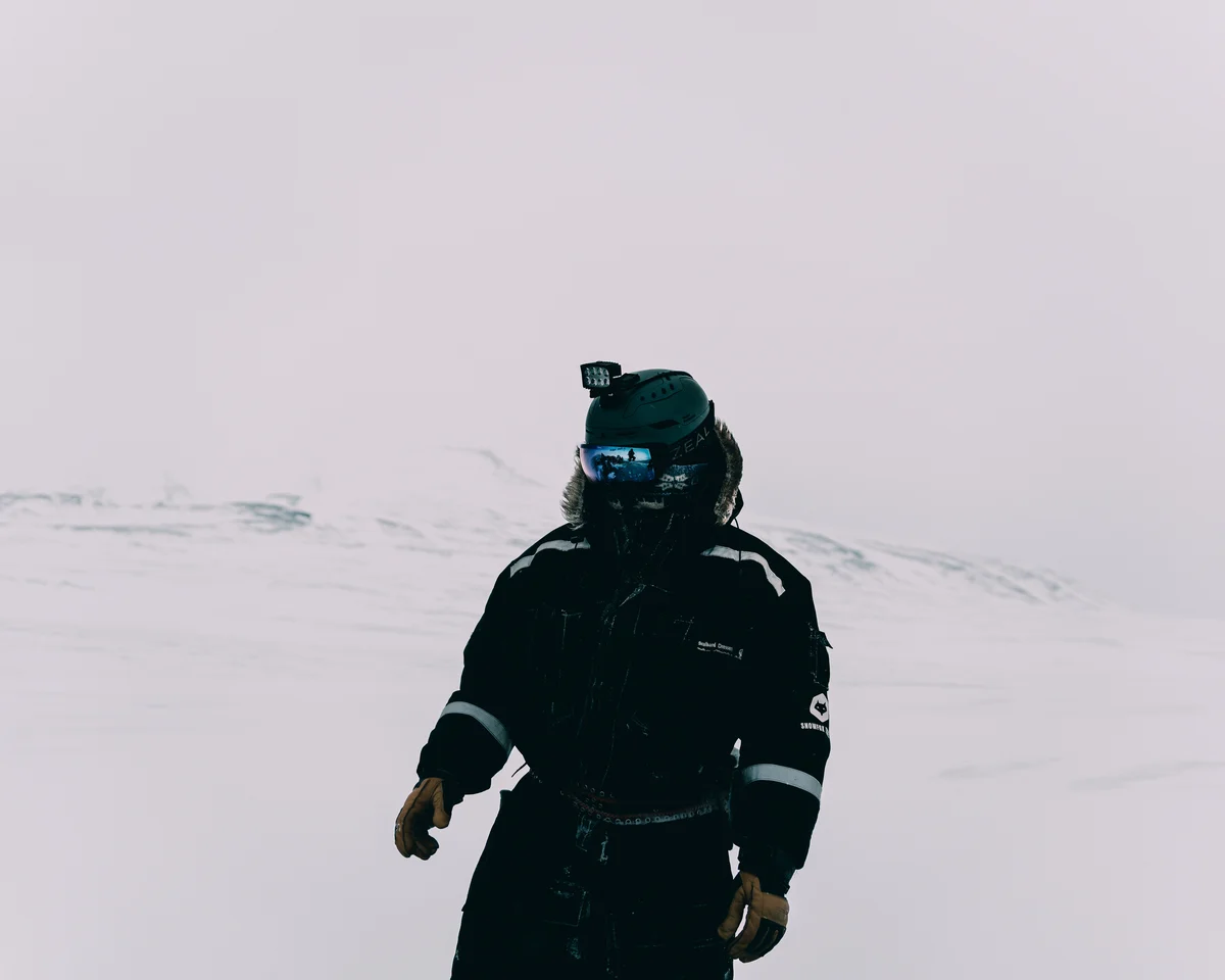A guest in a black expedition suit and helmet alone on an empty white plain, the rest of the group reflected small in the mirrored goggles
