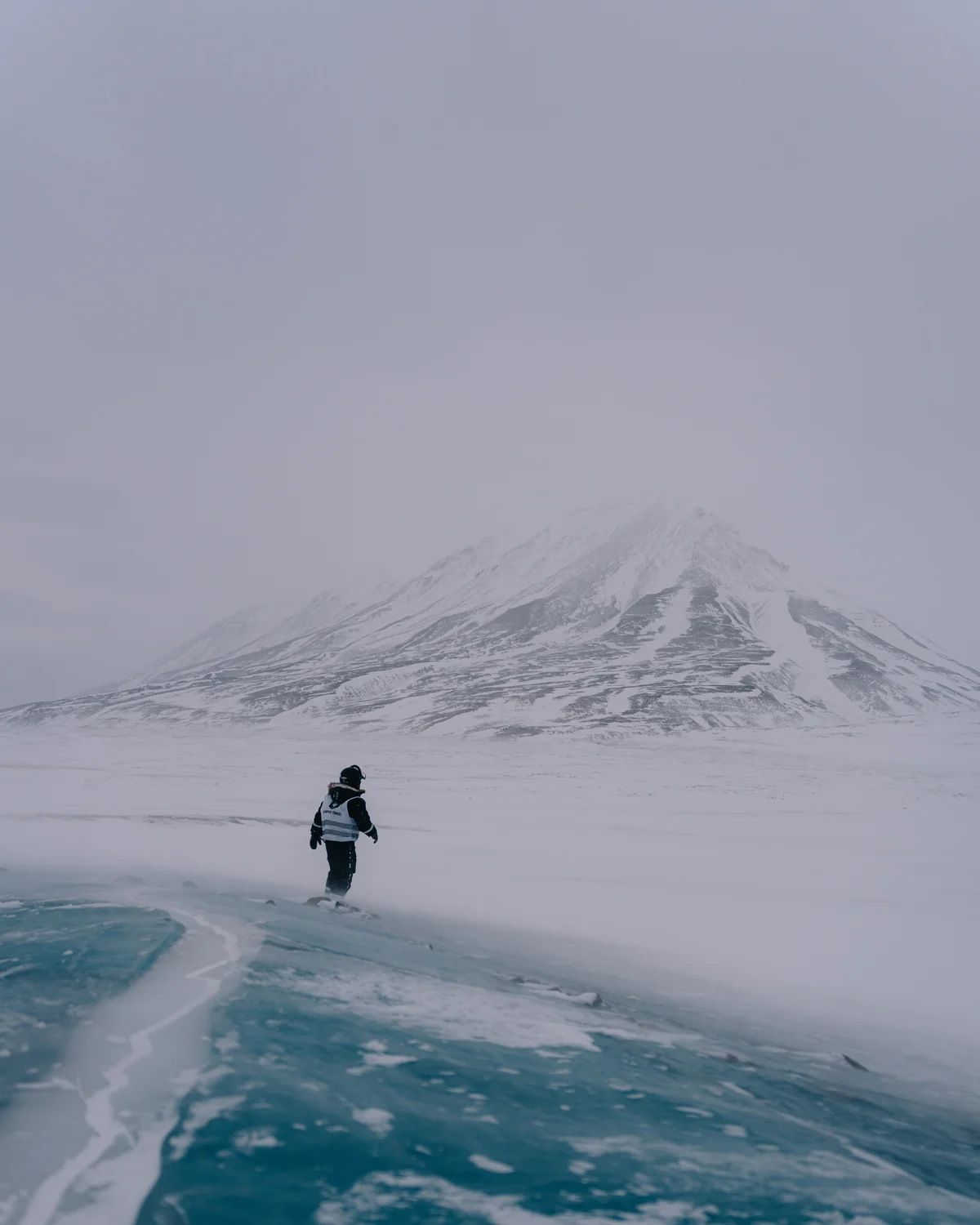 A lone figure in a high-visibility vest standing on bare turquoise lake ice, drifting snow crossing the foot of a snow mountain behind