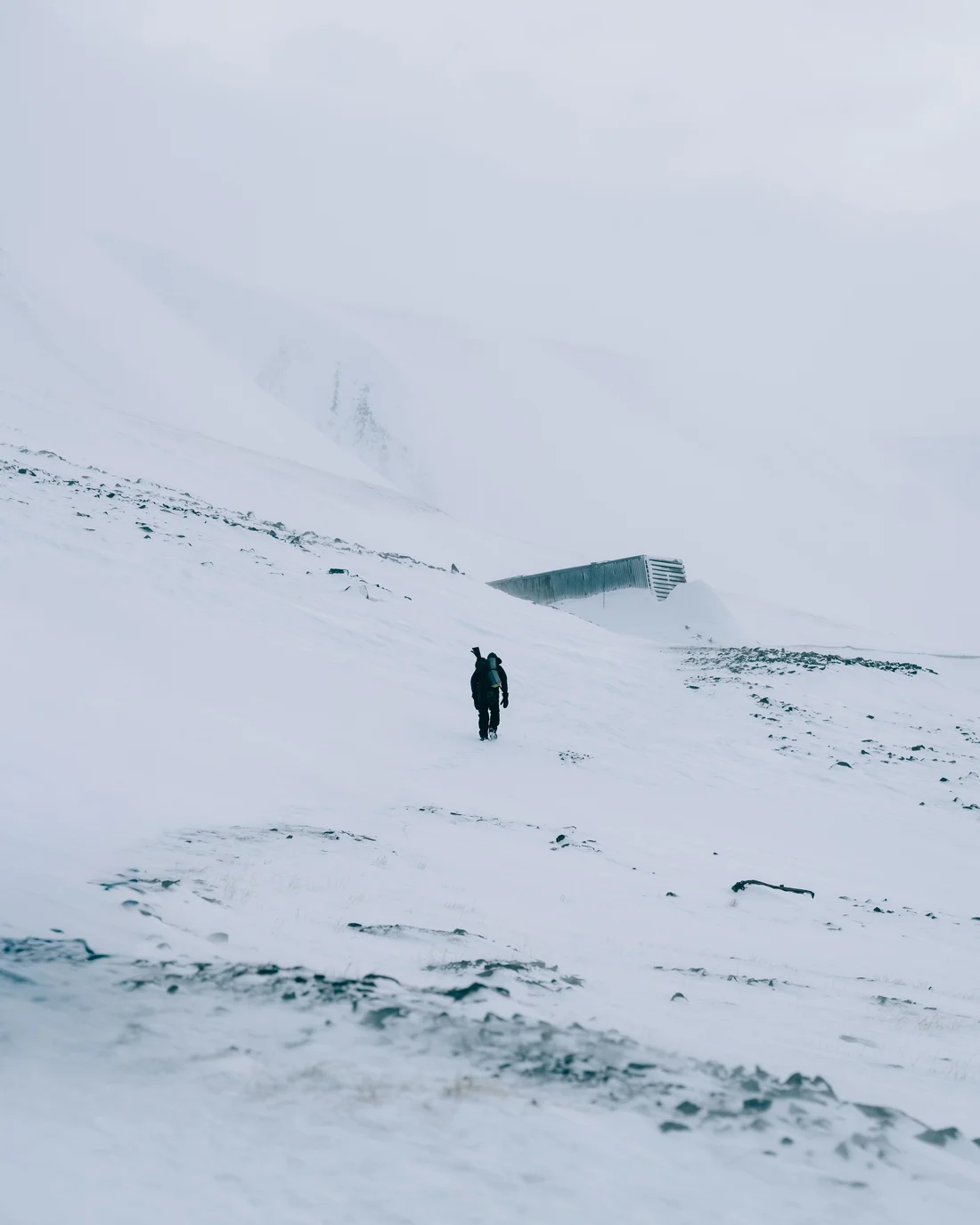 A single walker crossing a wide snow slope toward the low dark entrance building set into the ridge above