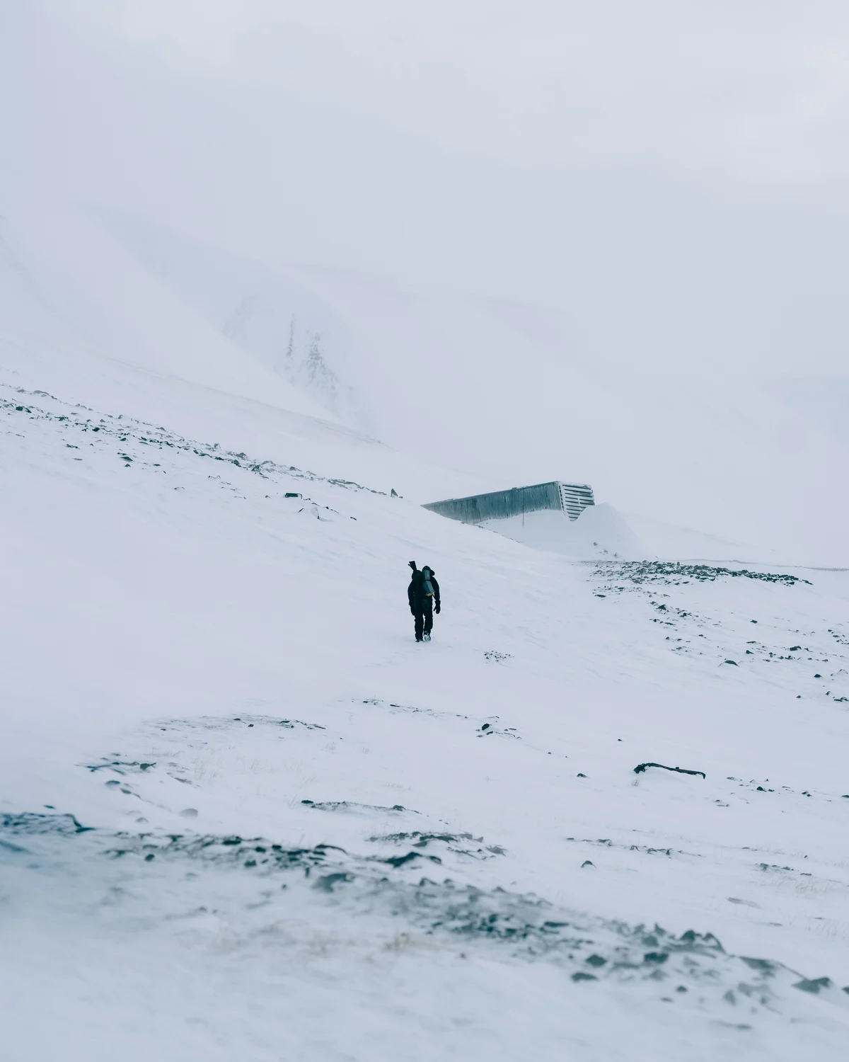 A single walker crossing a wide snow slope toward the low dark entrance building set into the ridge above