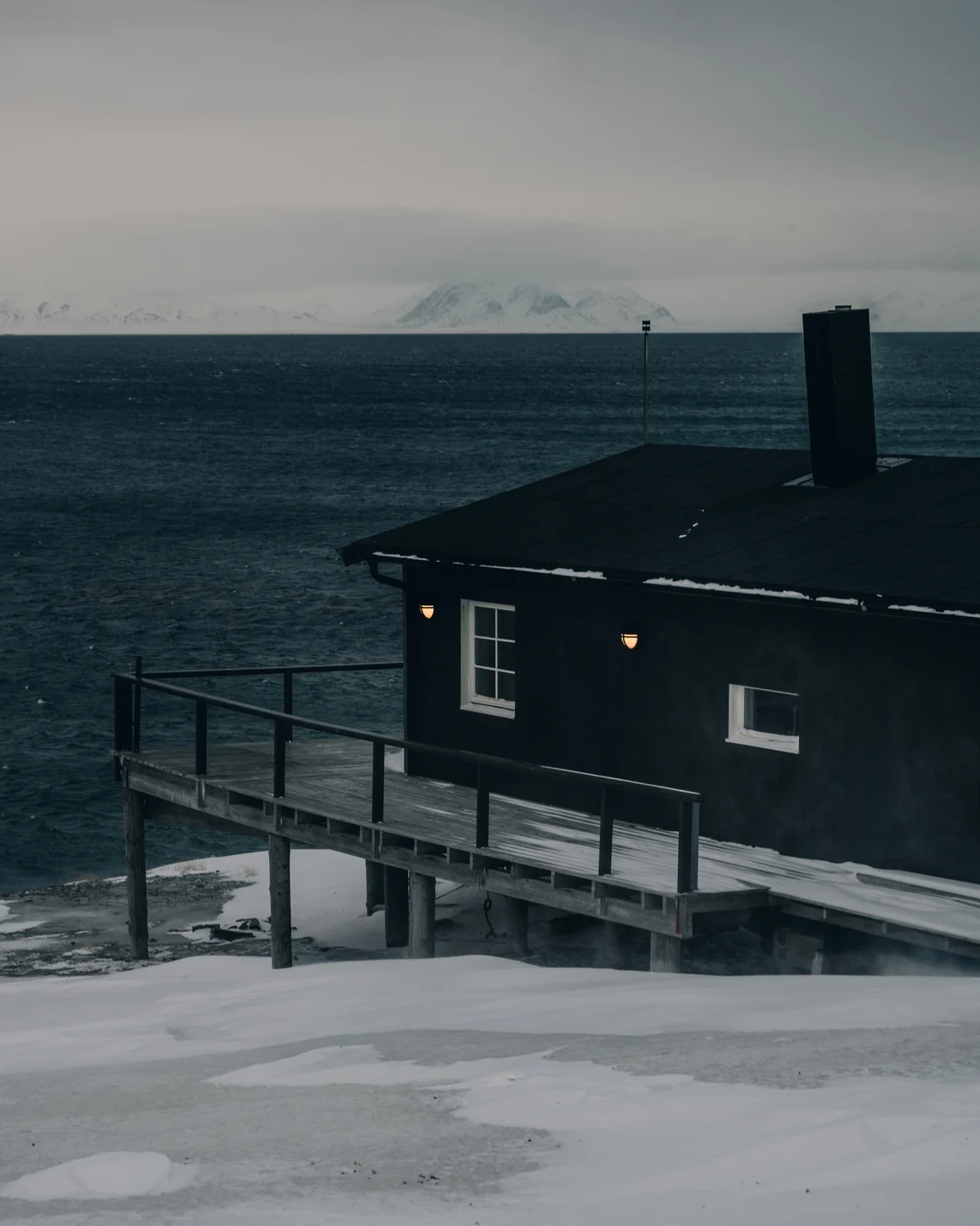 A black timber cabin on stilts at the water edge, two warm wall lamps burning against the dark sea and a snow range on the far shore