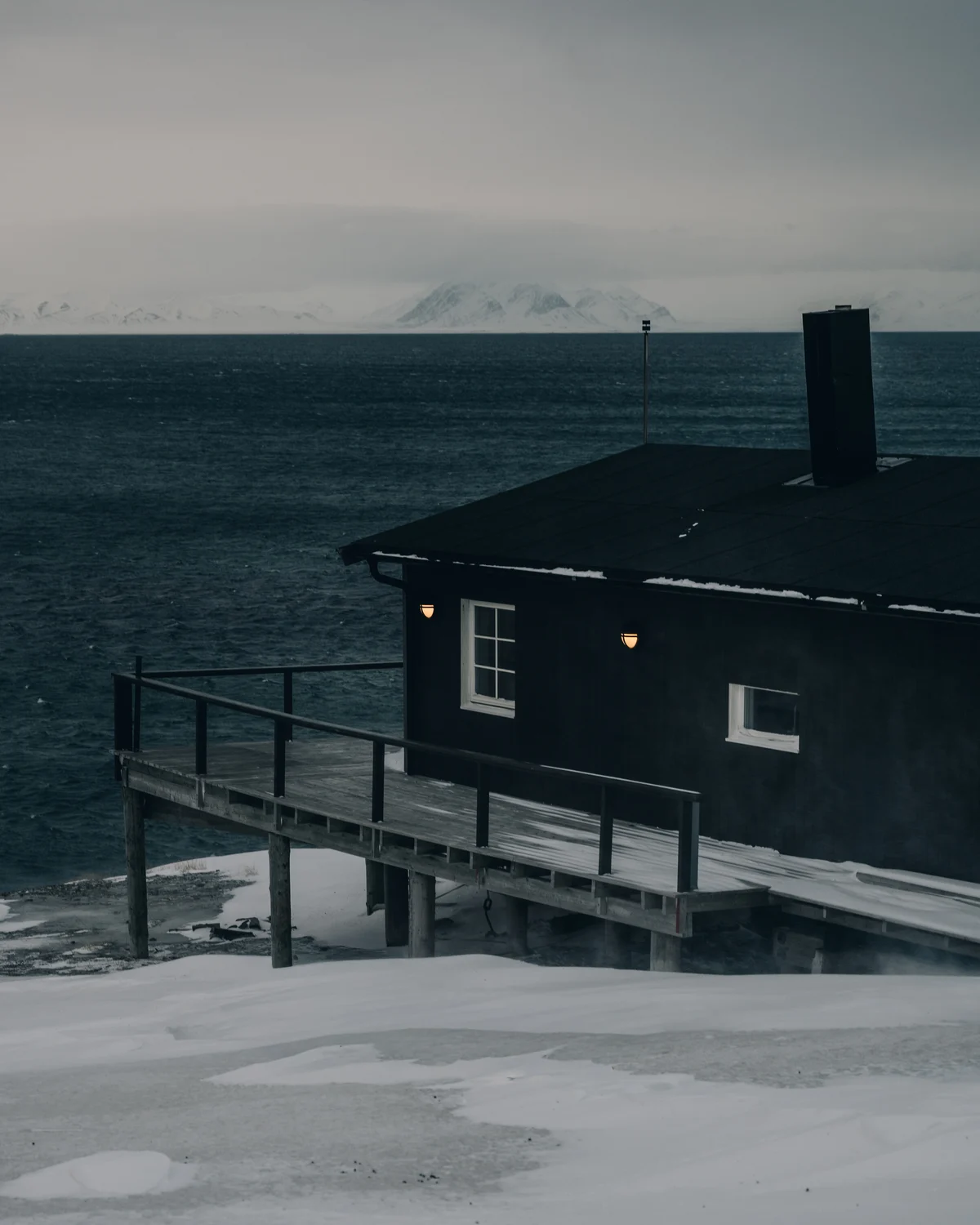 A black timber cabin on stilts at the water edge, two warm wall lamps burning against the dark sea and a snow range on the far shore