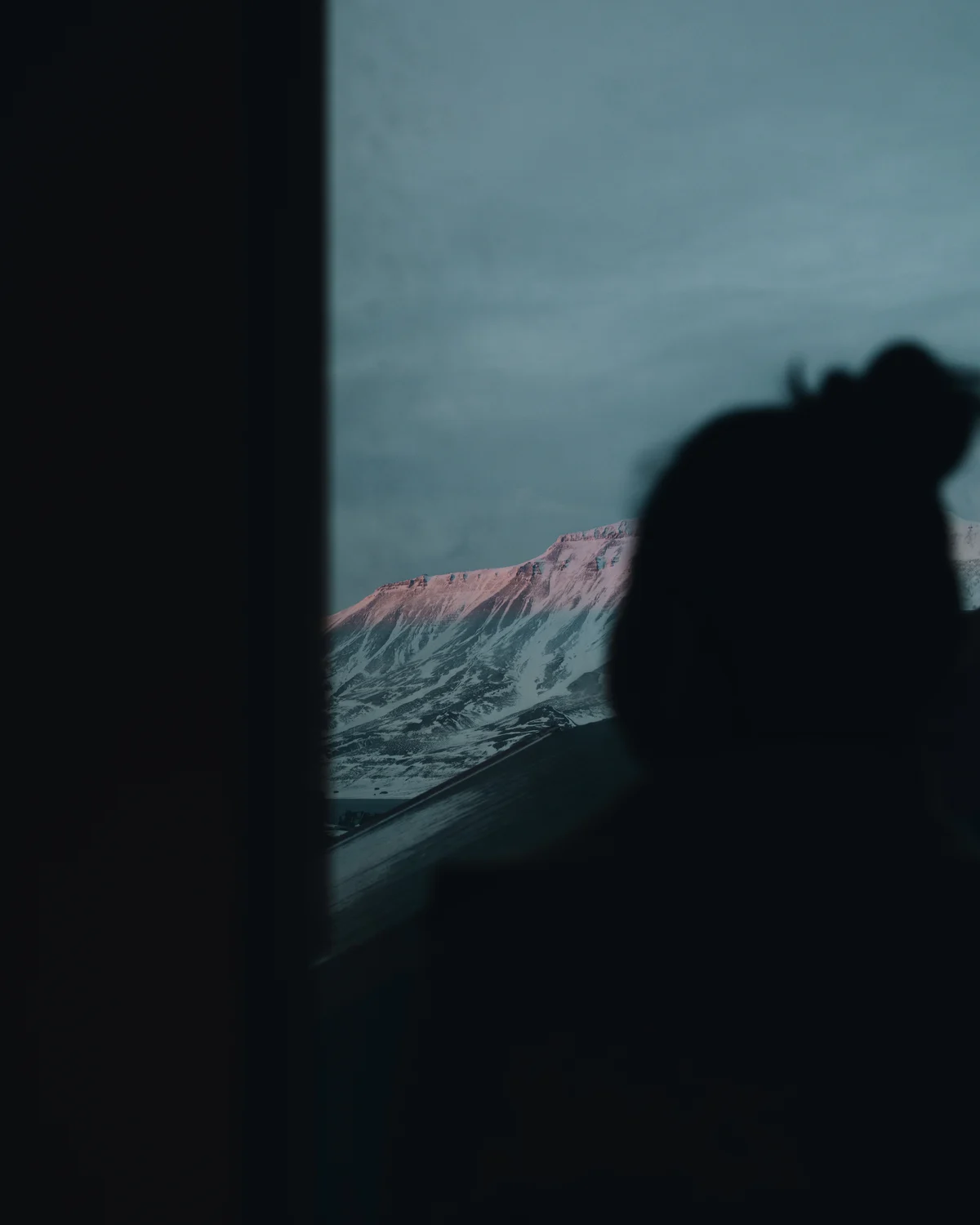 Someone silhouetted in a dark window against a flat-topped mountain, its summit catching the last pink light