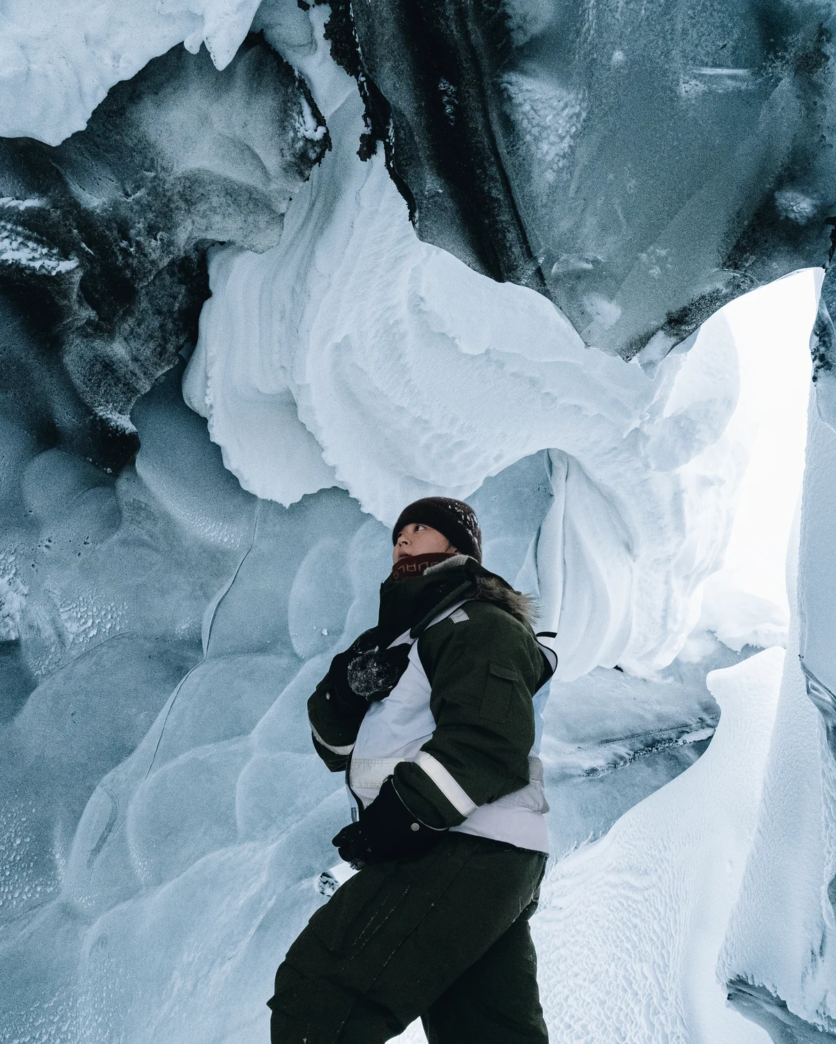 A figure in a dark parka standing inside a glacier cave, sculpted walls of blue and white ice curling overhead