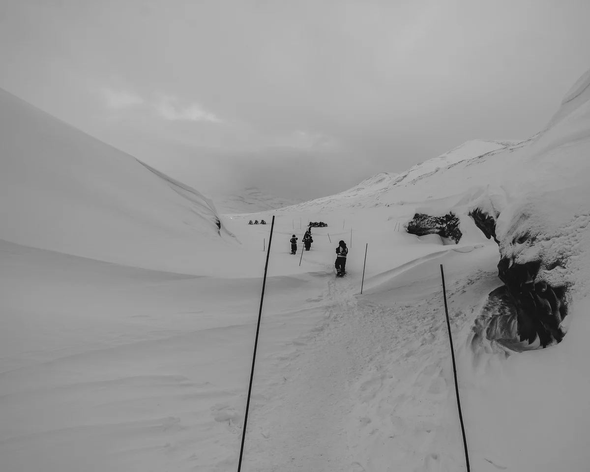 A line of people walking a marked trail up a broad snow valley, a torn blue opening in the glacier at the right