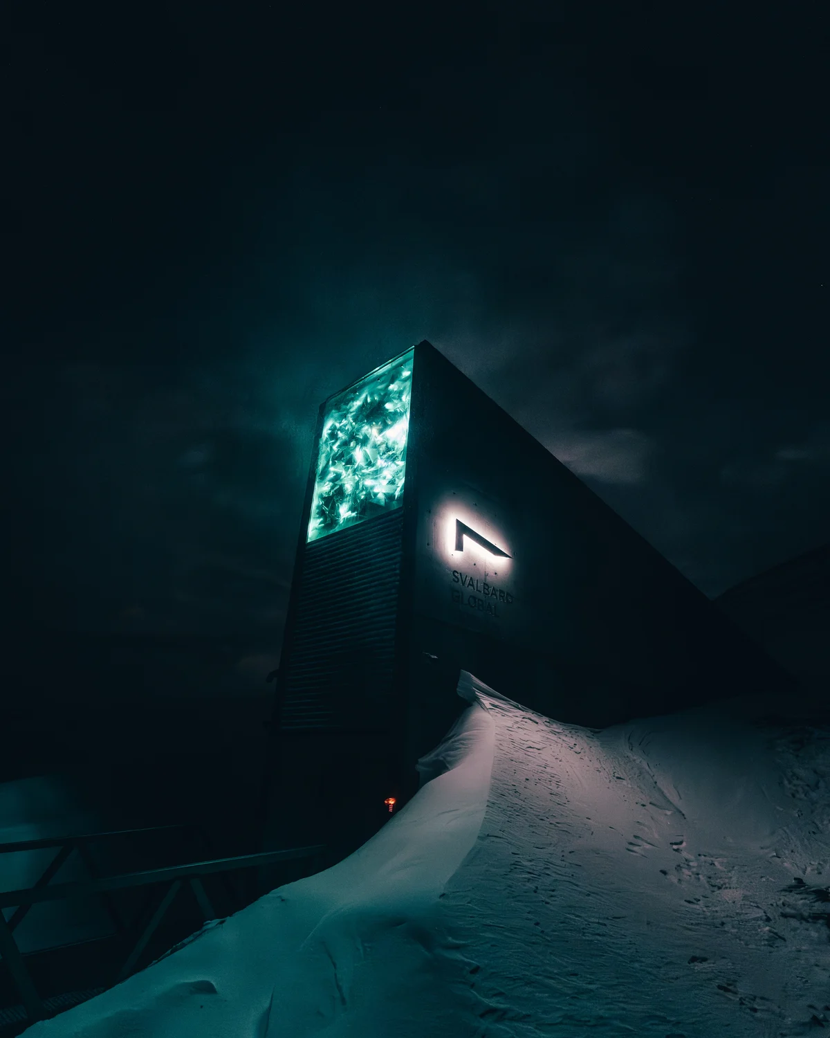 The concrete wedge of the Global Seed Vault at night, its face lit from within in green and white, snow drifted up against the door