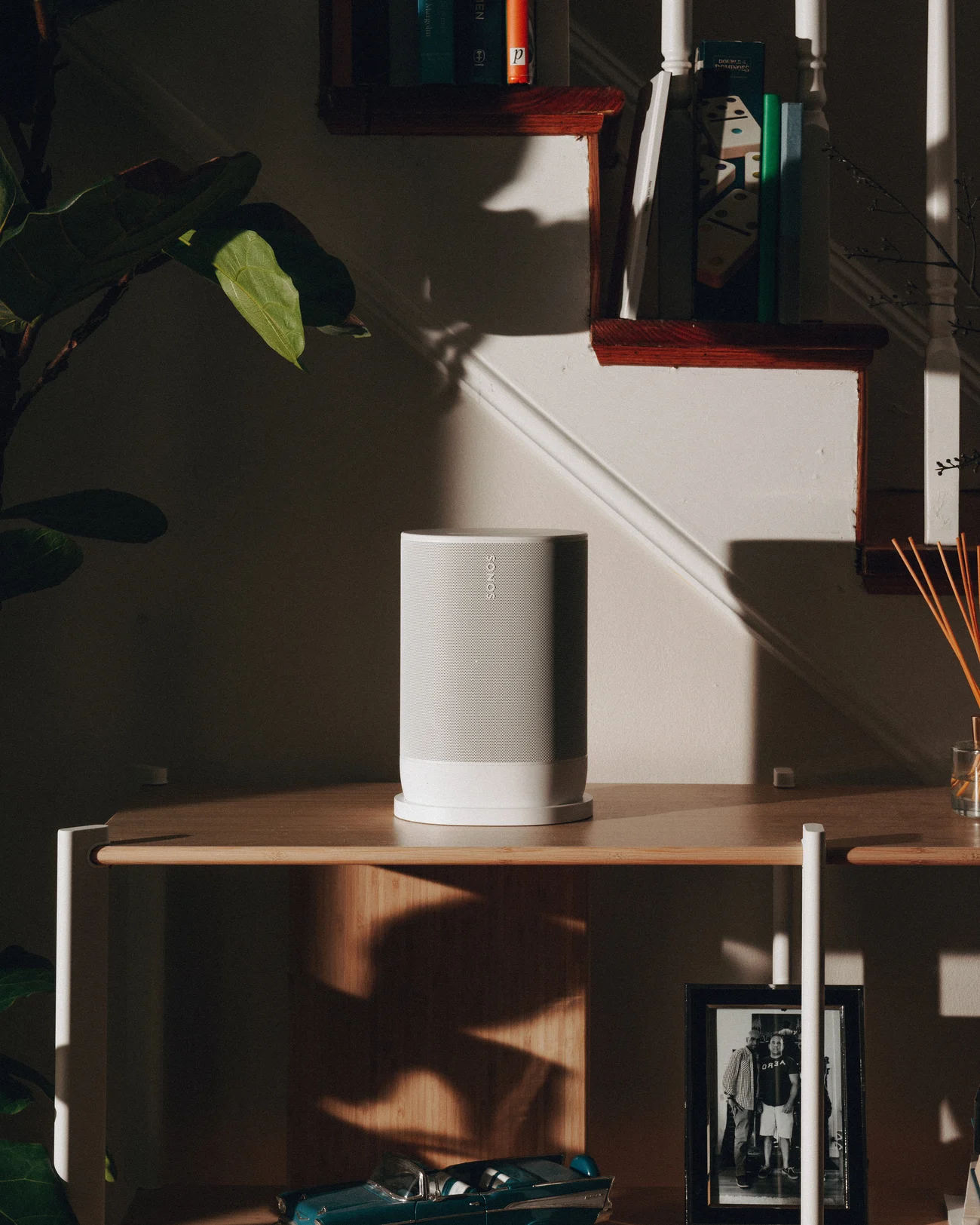 A white portable speaker on its charging base on a bamboo console, in a shaft of afternoon light, with a fiddle leaf fig and a staircase behind it