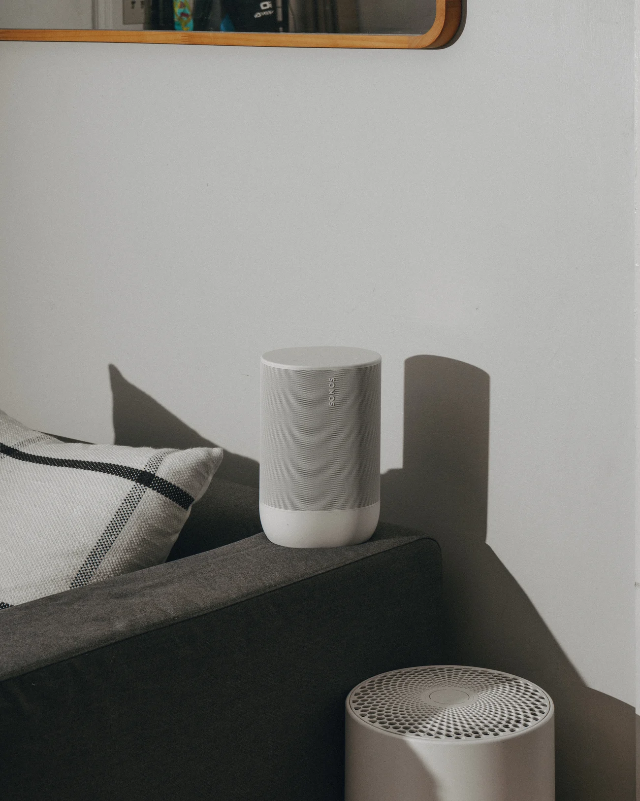 A white speaker on the arm of a dark grey sofa beside a striped cushion, casting a hard shadow on the wall, with a second speaker in the foreground