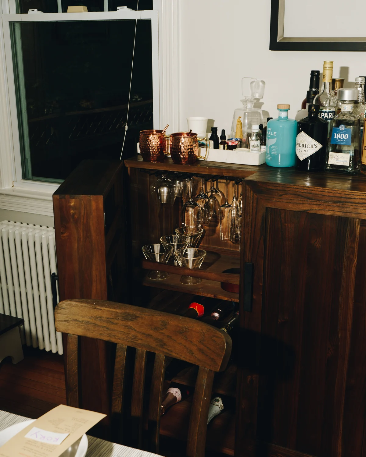 A home bar cabinet open to show hanging wine glasses and coupes, with copper mugs, gin, tequila and Campari on the top beside a laid table