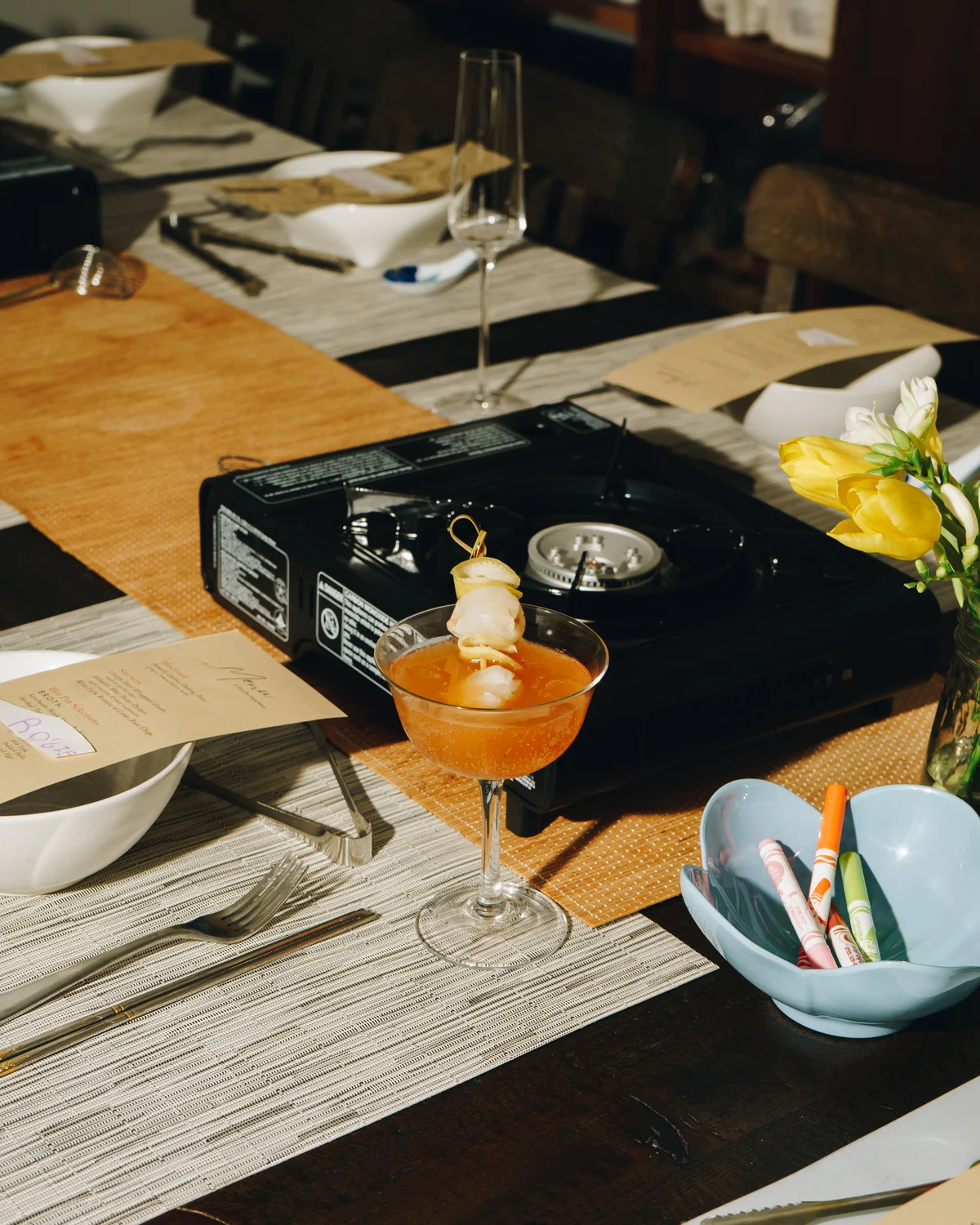 An amber cocktail in a coupe garnished with a skewer of prawn and lemon, on a laid table beside a portable gas burner, a handwritten menu and yellow tulips