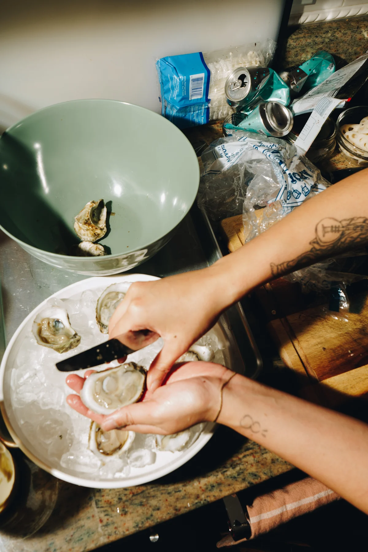 A chef shucking oysters over a plate of ice at a home kitchen counter, an oyster knife in one hand and the opened shell in the other