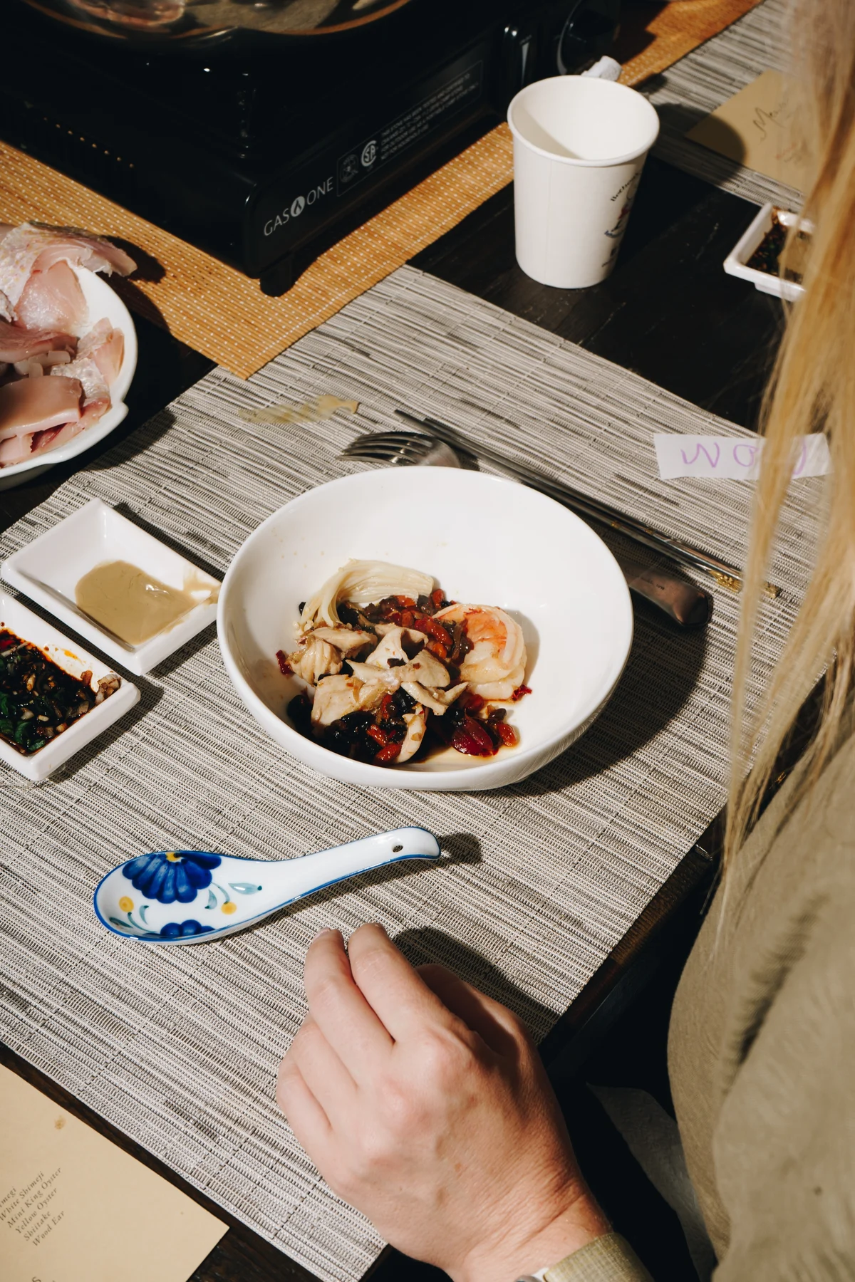 A shallow bowl of poached seafood with black beans and chilli on a woven placemat, beside two dipping sauces and a painted porcelain spoon