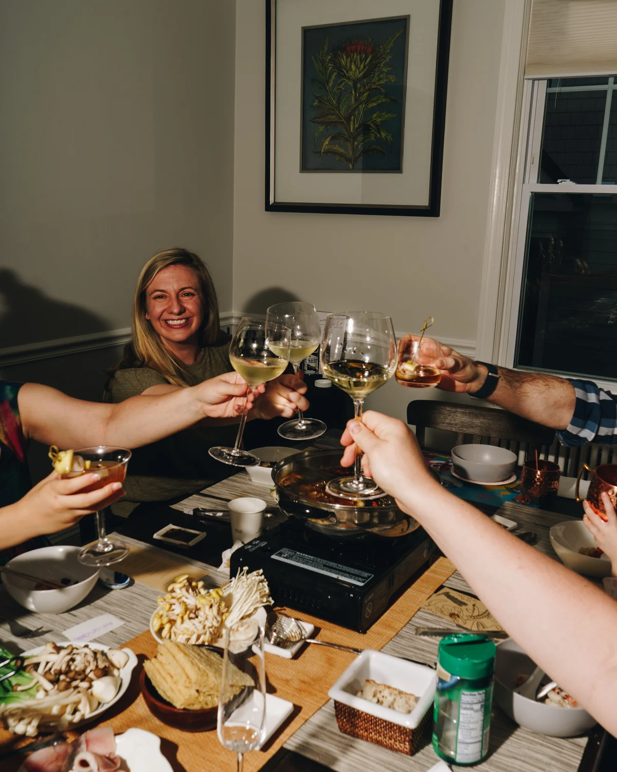 Five people reaching in to clink glasses over a bubbling hotpot on a portable burner, a woman smiling at the head of the table