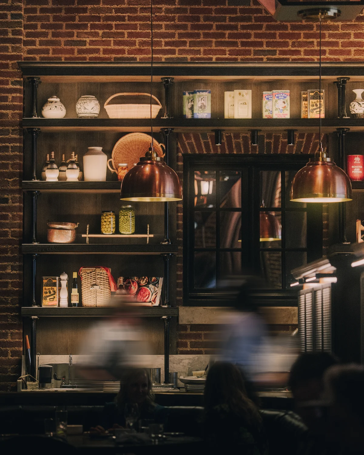 Shelves of olive oil tins and preserving jars lit under two copper pendant lamps on a brick wall, with diners passing in front of them in a blur