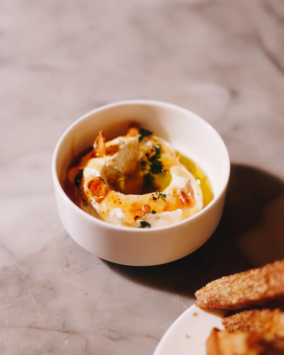 A bowl of whipped ricotta with olive oil, honey, herbs and crisped garlic, on marble beside a plate of sesame toasts