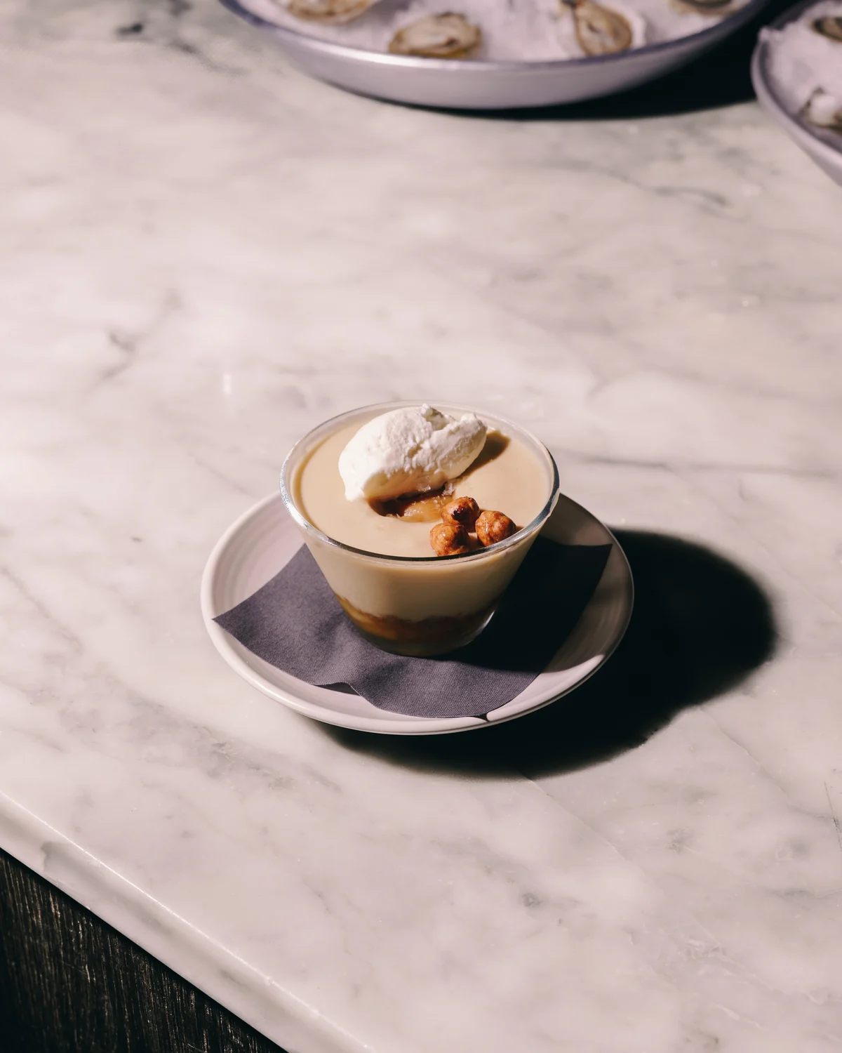 A glass of butterscotch pudding with a spoonful of cream and three glazed hazelnuts, on a saucer on a marble counter