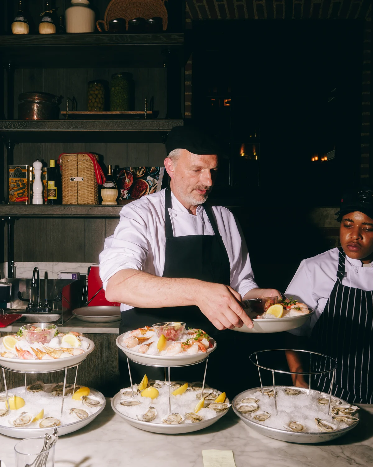 Two chefs at a marble raw bar building tiered seafood stands, plates of oysters on ice laid out in front of them and shelves of preserves behind