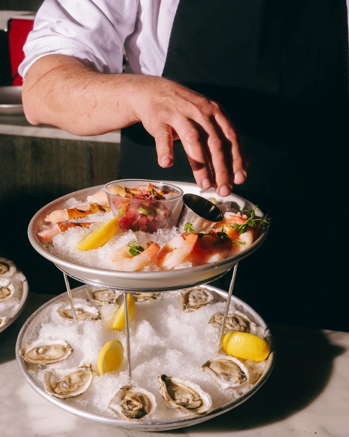 A chef reaching over a two tier seafood stand of shrimp, crab, tuna tartare and lemon on the upper plate and oysters on ice below