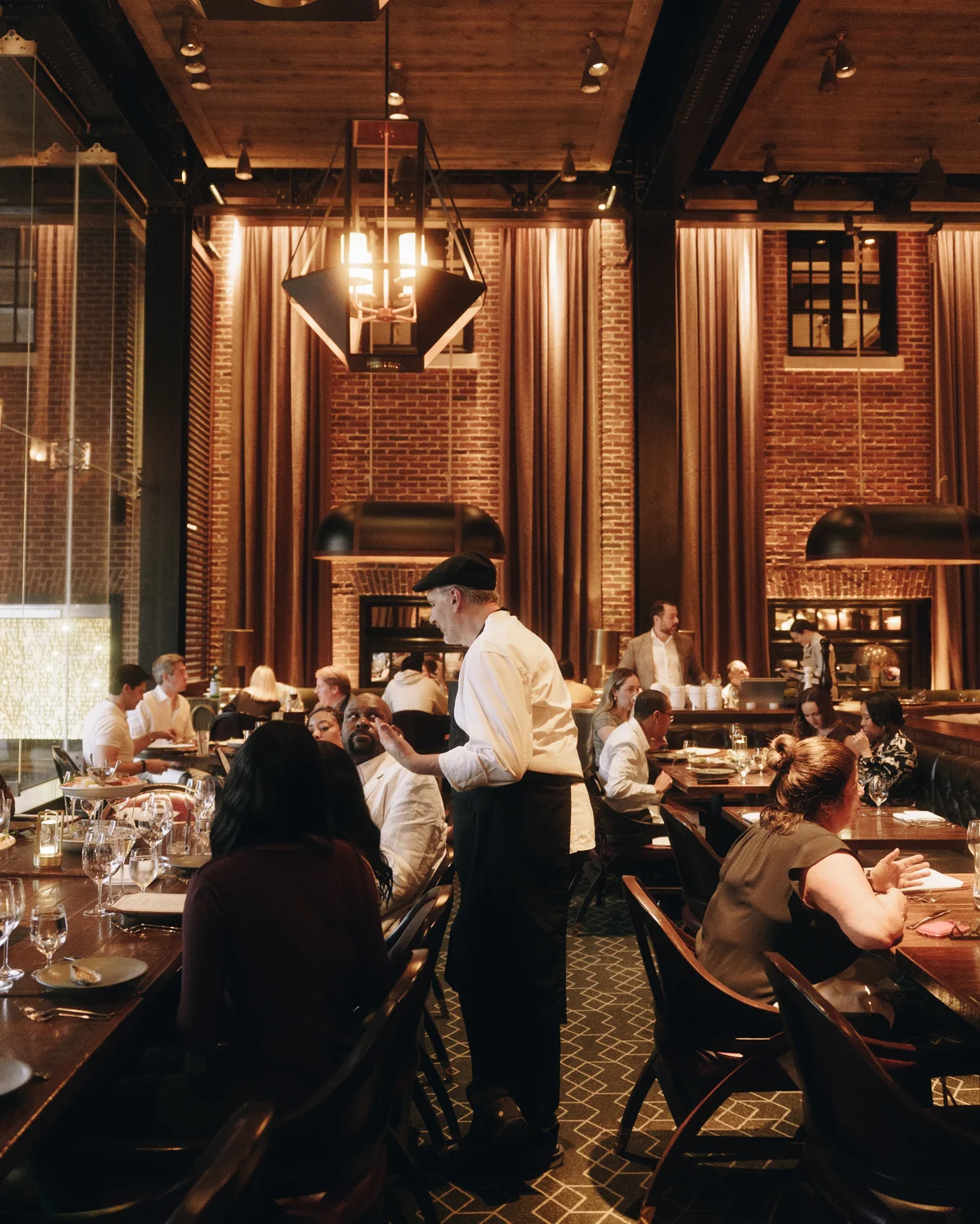 A busy dining room with double height brick walls, tall curtains and a lantern pendant, a waiter in a flat cap standing to talk to a table of four