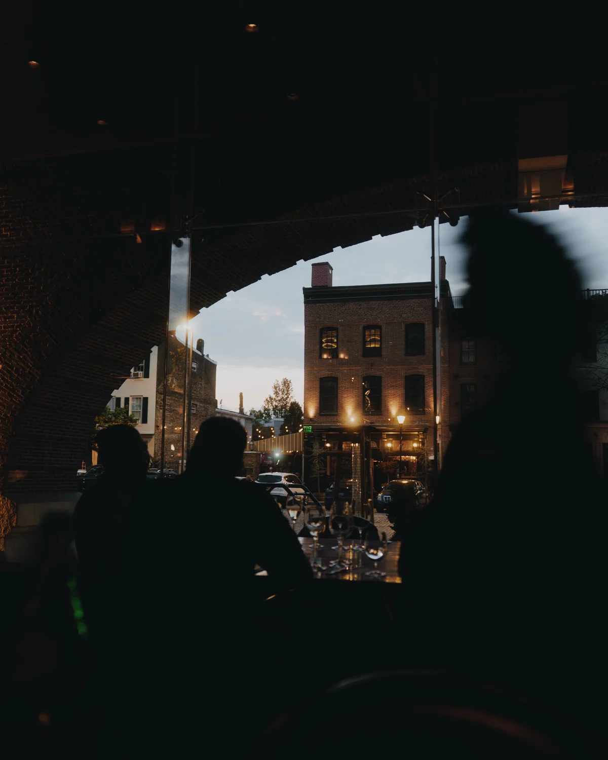 The view out of an arched restaurant window at dusk, brick warehouses and street lamps across the road, diners in silhouette at the table