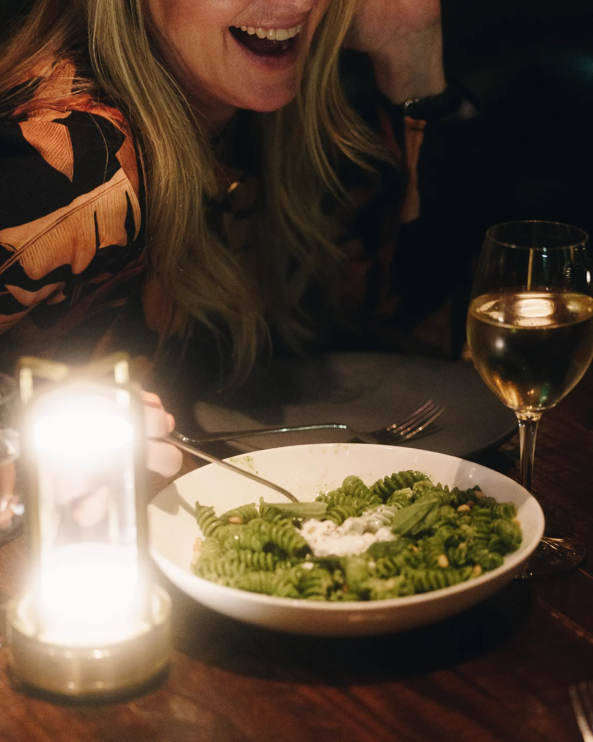 A bowl of green pesto fusilli with pine nuts and a spoonful of ricotta, on a candlelit table, with a woman laughing behind it