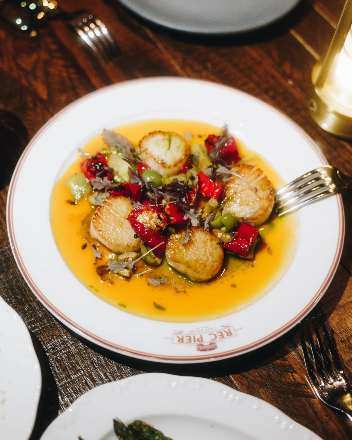 Four seared scallops in a saffron broth with red peppers, olives and purple shiso on a Rec Pier plate, a fork resting in it