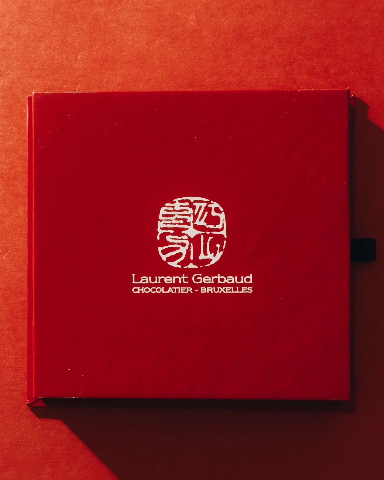 A red chocolate box closed on a red background, its lid stamped with the chocolatier seal and the words Chocolatier Bruxelles