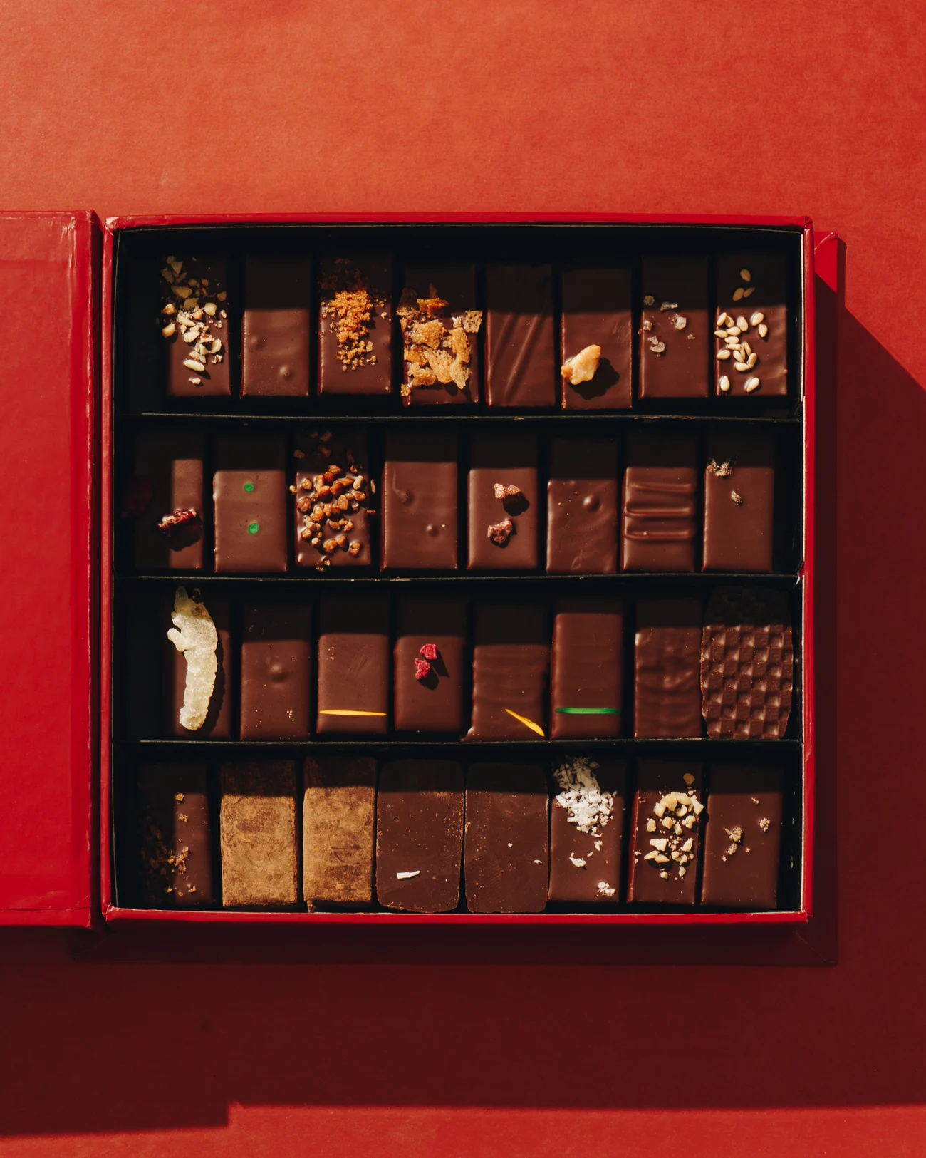 A close crop of four rows of chocolates in a box, each row finished differently: caramelised crumbs, chopped hazelnuts, dried raspberry and coloured piping