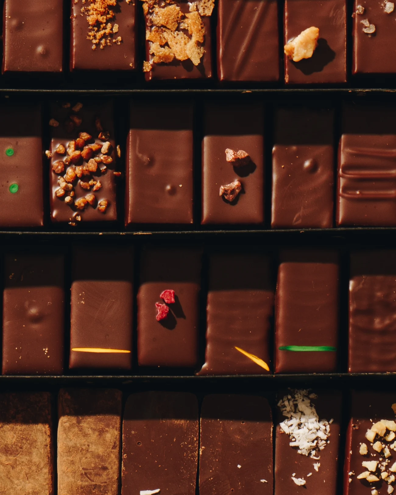 An open box of chocolates from above, four rows of rectangular pralines finished with candied peel, nuts, coconut, dried fruit and coloured lines