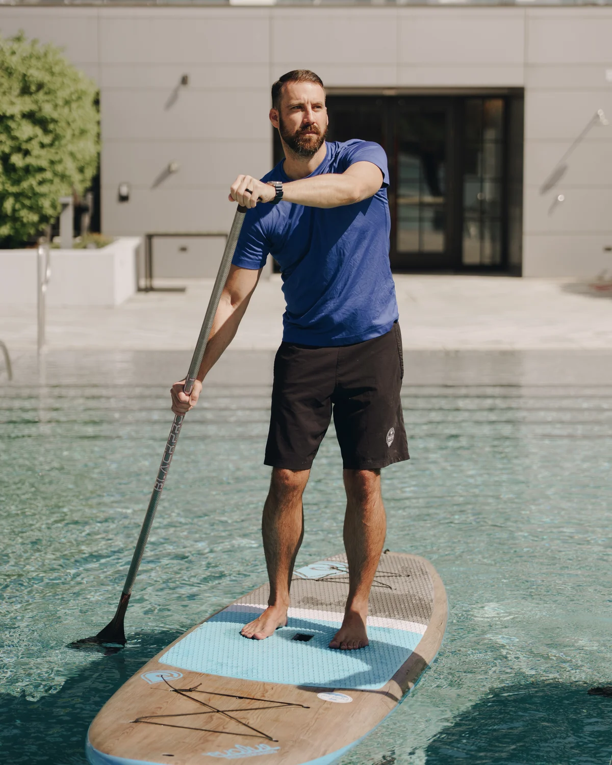 A man standing on a paddleboard mid-stroke, paddle planted in the pool