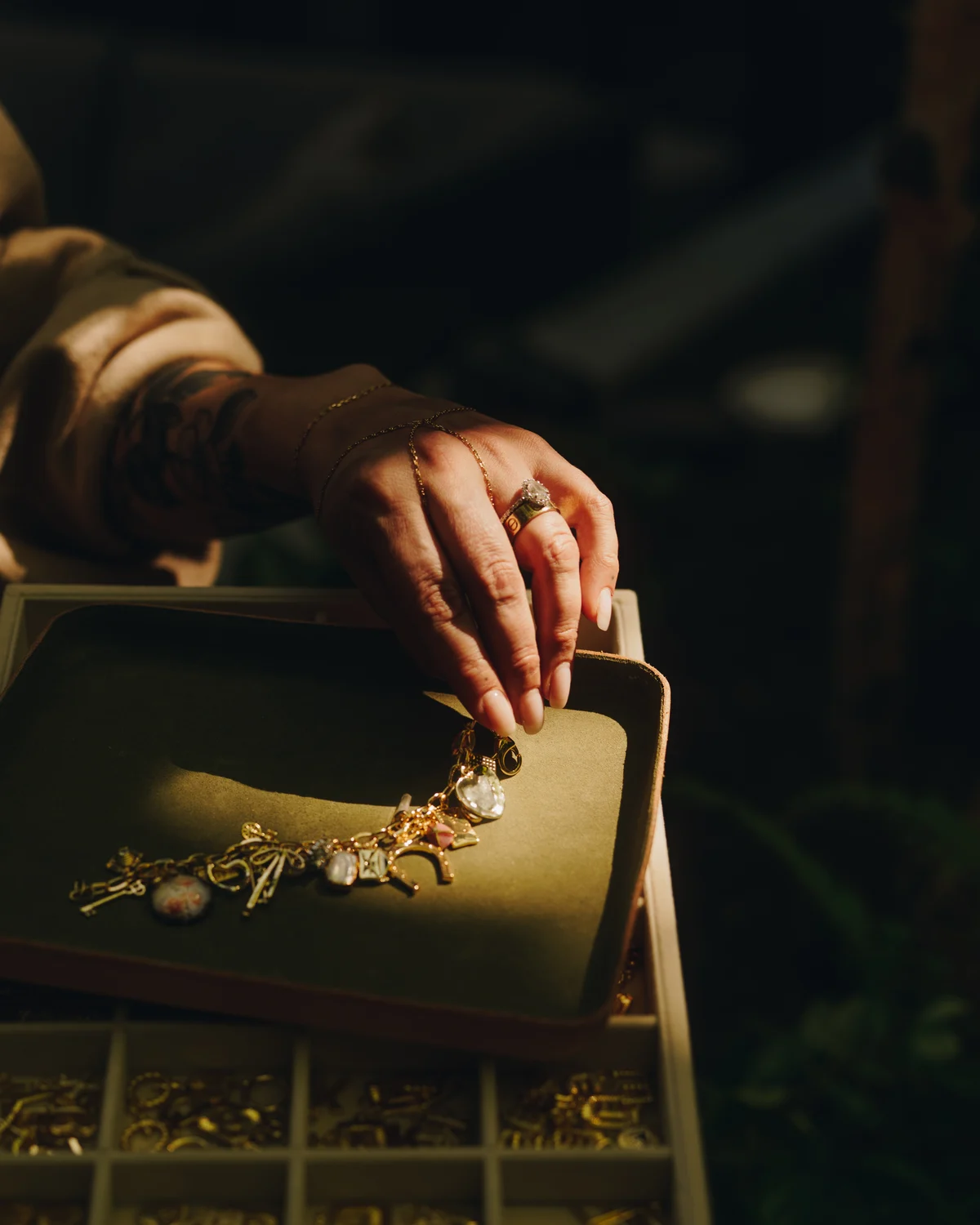 A hand reaching into a lined tray of gold charms, a compartmented case of more of them below