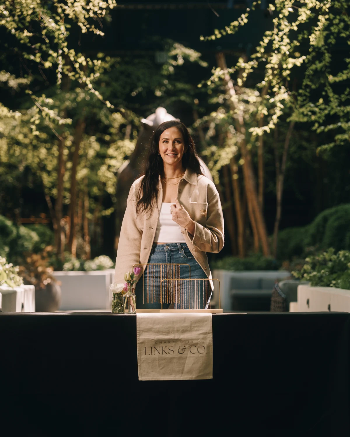 The owner standing behind her table under the trees at golden hour, a canvas runner printed with the shop name hanging over the front