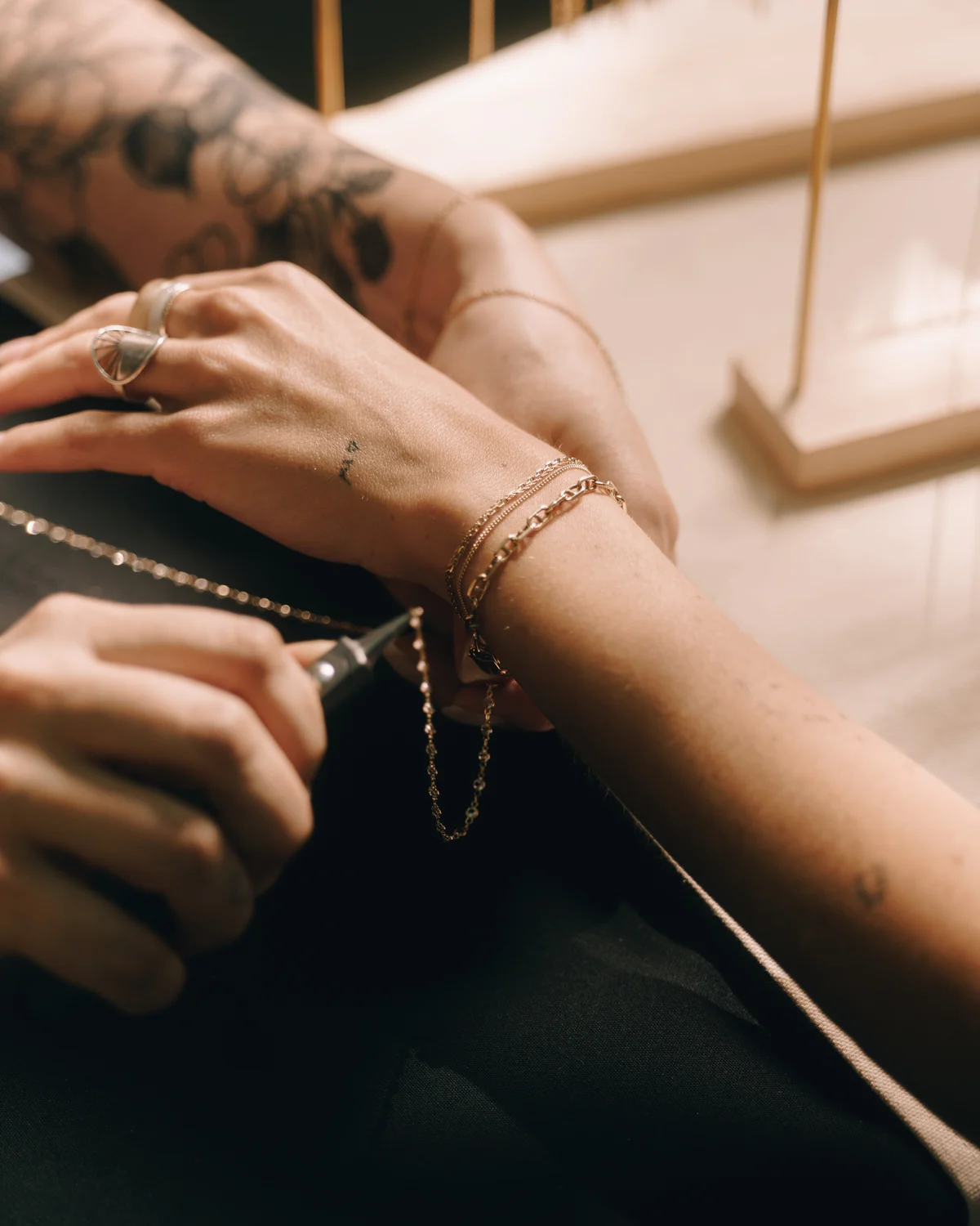 Hands fastening a fine chain around another person's wrist, rings and stacked bracelets close in the frame