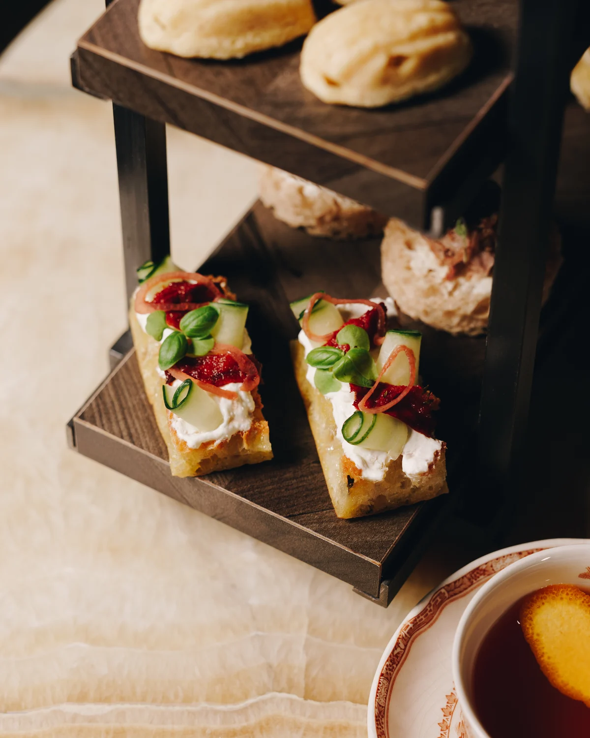 Two pieces of focaccia with cream cheese, cucumber ribbons, pickled onion and basil on a tea stand, with scones on the tier above