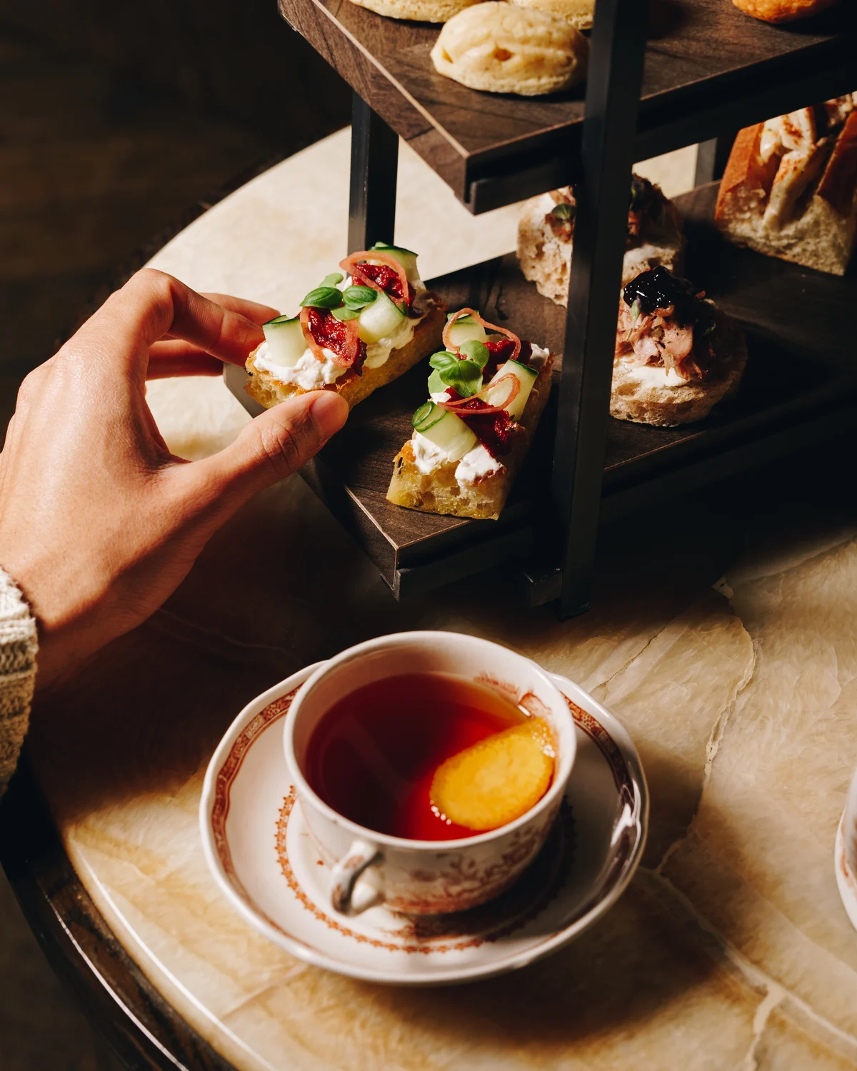 A hand holding a slice of focaccia topped with cream cheese, cucumber, pickled onion and basil, above a cup of tea with an orange slice in it