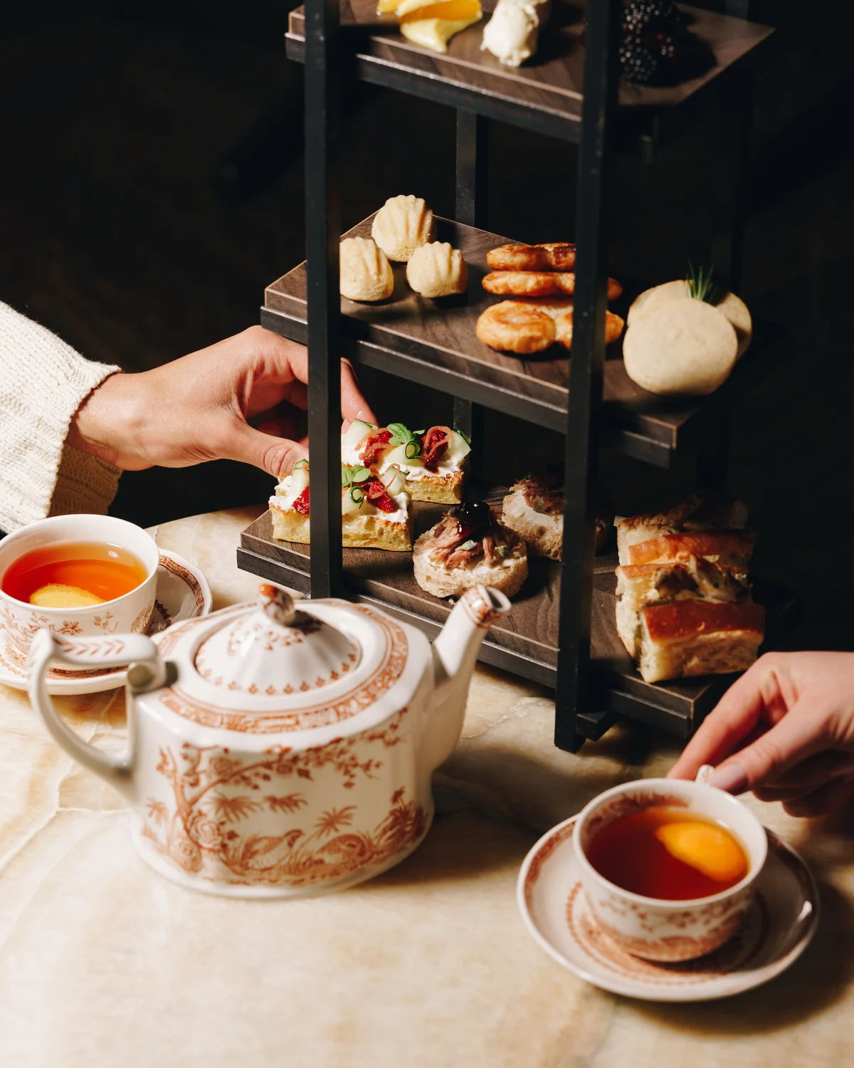 A hand lifting a canape from the middle tier of an afternoon tea stand, a second hand reaching for a teacup on the other side of the teapot